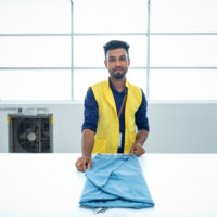 man standing in front of the bright window of a textile factory folds a blue garment and smiles at the camera