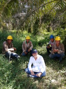 a woman smiling with three men wearing construction helmets surrounded by oil palm trees