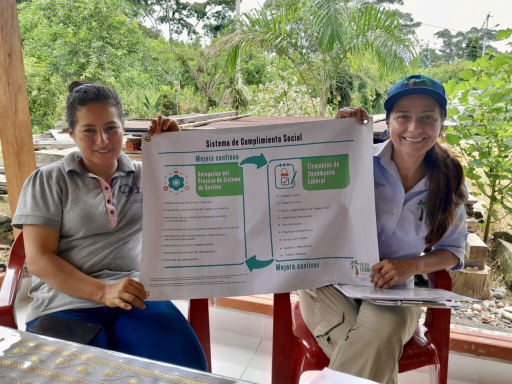 Yolanda and a woman smile while holding either side of a poster describing social compliance systems